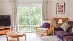 Un salon comprenant un téléviseur, une table ronde en bois, un canapé violet, un pouf à motifs, un fauteuil en bois avec un coussin violet et une vue sur la verdure grâce à de grandes fenêtres.