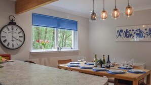 Une cuisine dotée d’un plan de travail en granit, de poutres apparentes en bois au plafond, d’une horloge, d’une fenêtre avec des stores bleus et d’un coin repas préparé pour un repas.