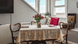 A dining area with a table covered by a lace tablecloth, two wooden chairs, a vase with flowers, and a red pillow on a bench. - 1 bedroom accommodation in Hastings (Hastings)