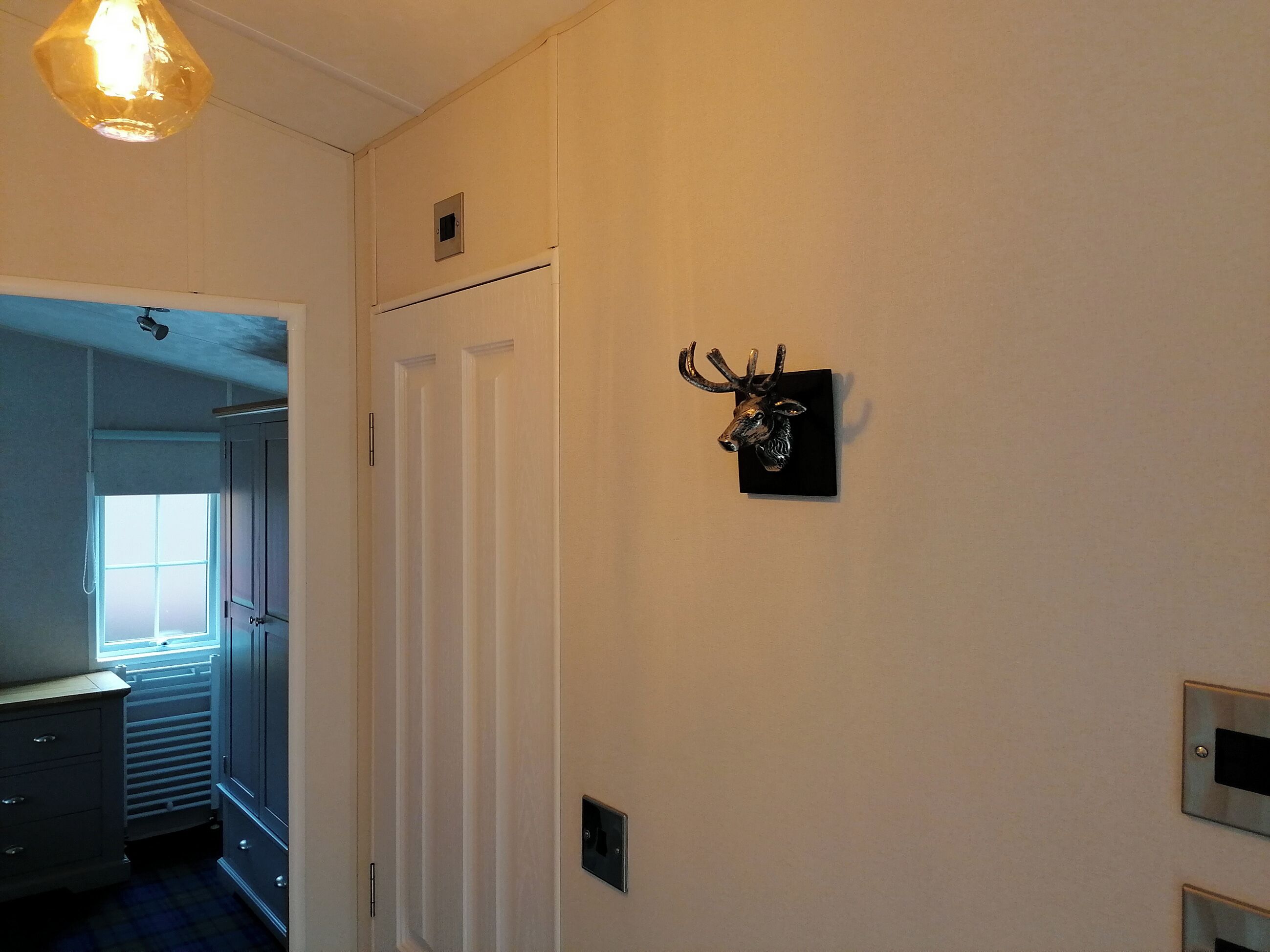 Un pasillo con un ciervo decorativo colgado en la pared, un radiador y una puerta que da a otra habitación.
