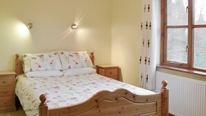 Un lit en bois avec une literie à motifs floraux, une table de chevet, une lampe fixée au mur et une fenêtre avec des rideaux.