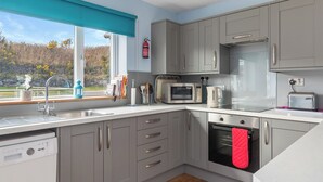 A modern kitchen with grey cabinets, a dishwasher, a microwave, a toaster, and a window with a view of trees. - Min y Traeth (By the Beach) (Morfa Nefyn, near LLyn Peninsula)