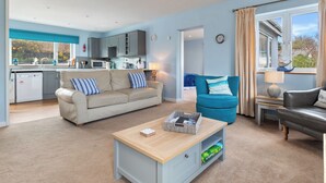 A modern living room with a sofa, a blue armchair, a coffee table, and a view of a kitchen and garden. - Min y Traeth (By the Beach) (Morfa Nefyn, near LLyn Peninsula)