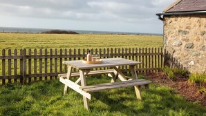 A picnic table with a bottle and glasses, set against a stone wall and a grassy field with a wooden fence. - 2 bedroom accommodation in Clynnog Fawr (Clynnog Fawr)