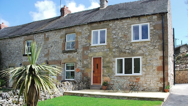 A stone house with a wooden door, multiple windows, and a small garden with a palm tree. - Godfrey Hole Cottage (Godfrey Hole near Carsington Water)
