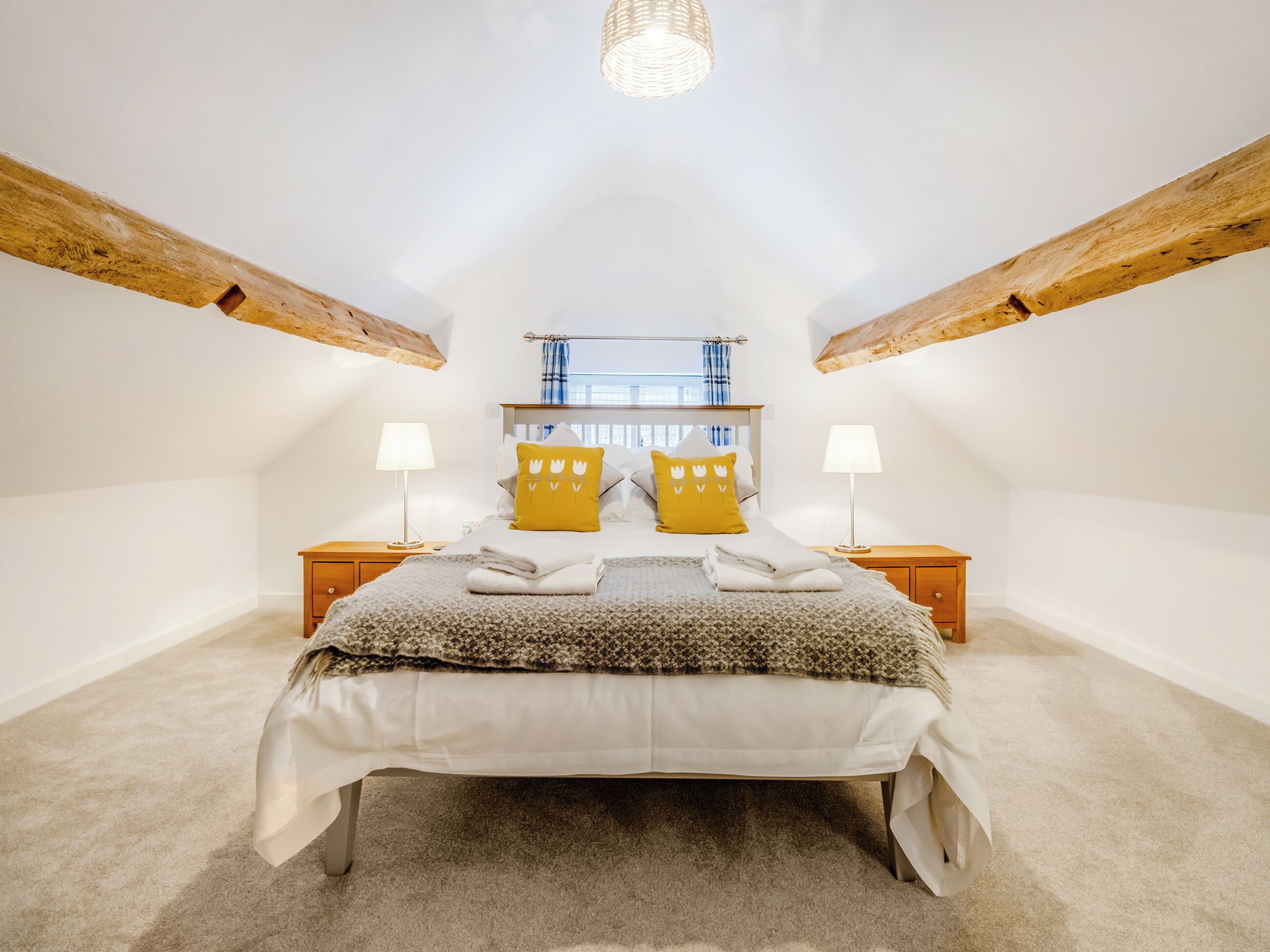 Une chambre avec un plafond en pente, des poutres en bois, un lit avec une literie blanche, deux tables de chevet avec des lampes et une fenêtre avec des rideaux bleus.