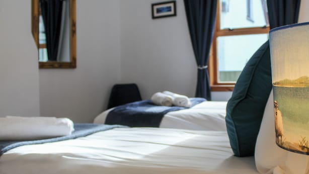 Una cama bien tendida con sábanas blancas, almohada azul oscuro y una lámpara con diseño de paisaje.