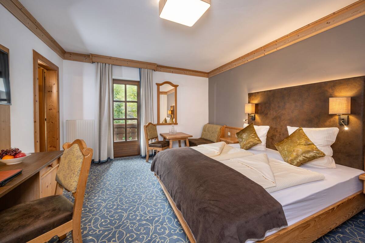 Ein Hotelzimmer mit einem großen Bett, einem Schreibtisch mit Stuhl, einem kleinen Tisch und einem Fenster mit Vorhängen.