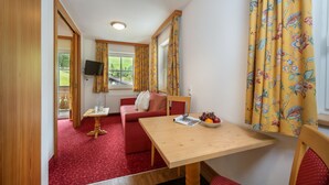 Une pièce avec une moquette rouge, une table en bois sur laquelle se trouve un saladier rempli de fruits, un canapé rouge et un téléviseur fixé au mur.