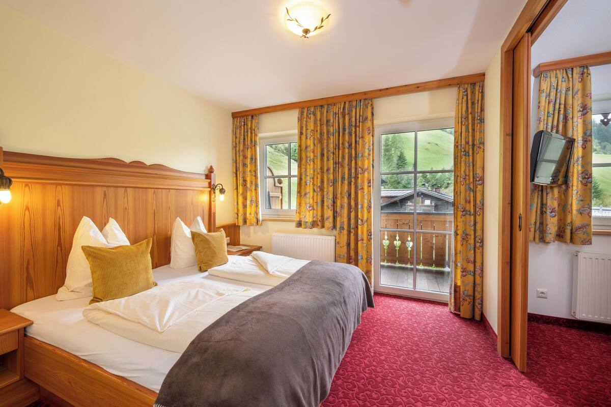 Ein Hotelzimmer mit einem großen Bett, einem Holz-Kopfteil und einem roten Teppich. Zwei Fenster mit Vorhängen, ein an der Wand montierter Fernseher und Blick auf ein Haus.