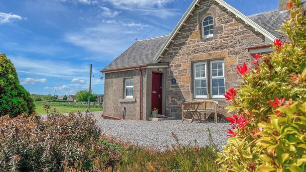 A stone house with a red door, a gravel driveway, and a garden with various plants. - Rowan Cottage - UK38991 (Dalcross)