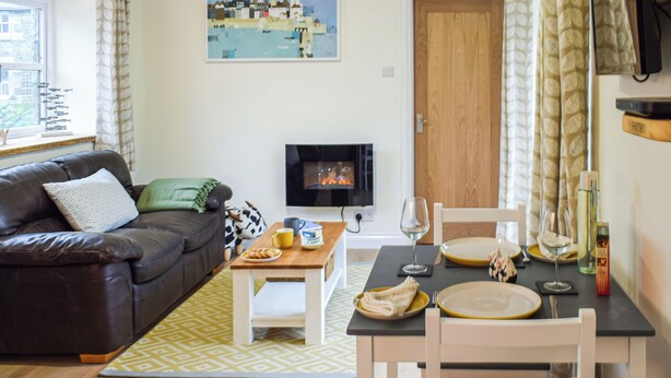 Una sala de estar acogedora con un sofá, una mesa de centro y una mesa de comedor lista para dos personas. Un televisor está montado en la pared, y se ve una ventana con cortinas.