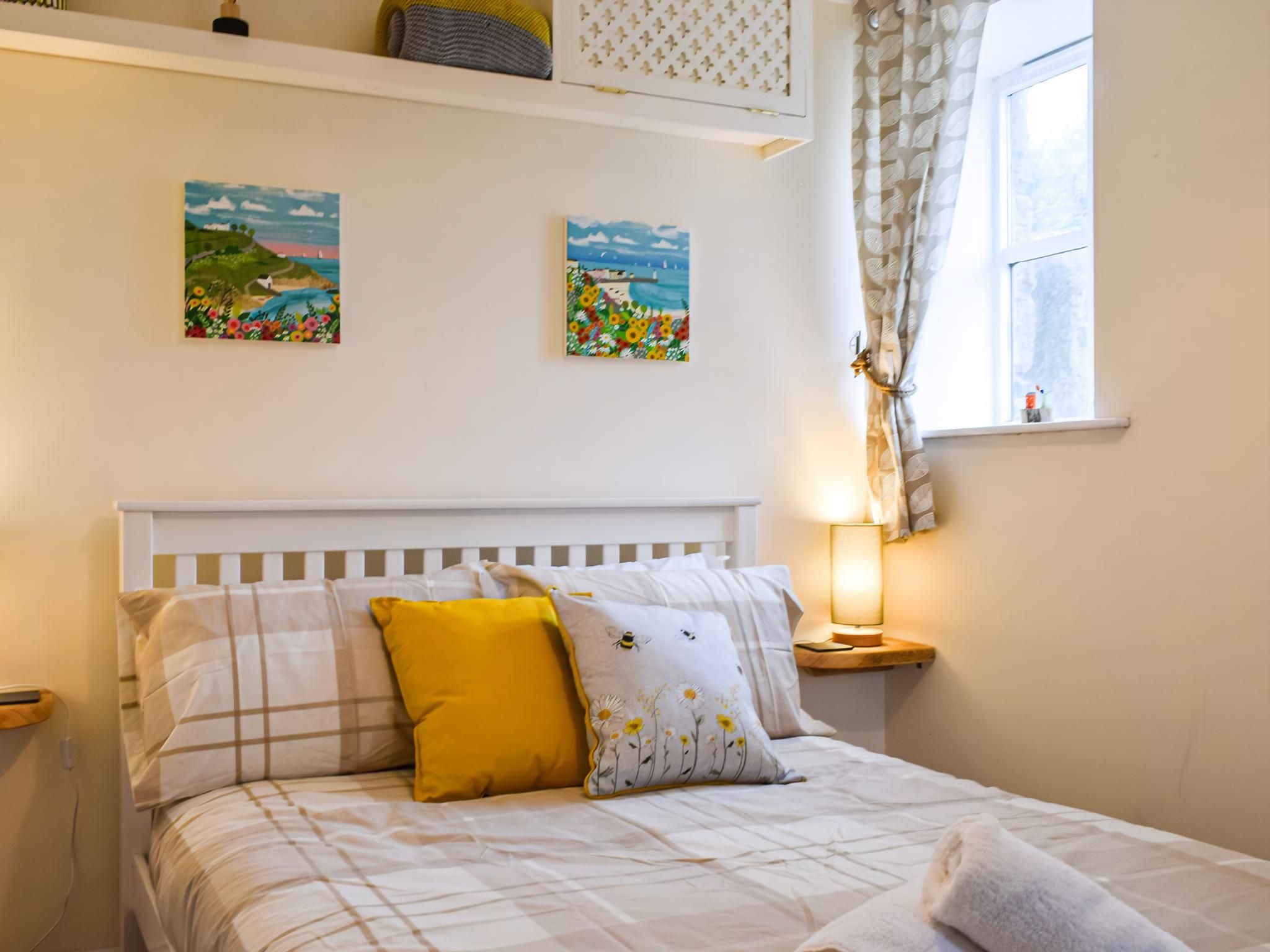 A neatly made bed with a checkered bedspread and a single yellow pillow, a bedside table with a lamp, and two wall-mounted paintings of landscapes.