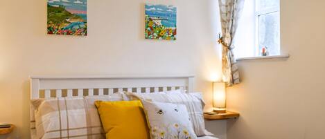A neatly made bed with a checkered bedspread and a single yellow pillow, a bedside table with a lamp, and two wall-mounted paintings of landscapes.