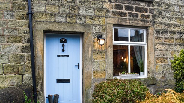 Un bâtiment en pierre avec une porte bleue portant l’inscription « 39 Shire Nook », une fenêtre à cadre blanc et un petit porche avec un paillasson et une paire de bottes noires.