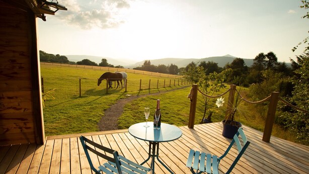 Une terrasse en bois avec une table et des chaises, un verre de vin et une vue sur un pré clôturé où paissent des chevaux.