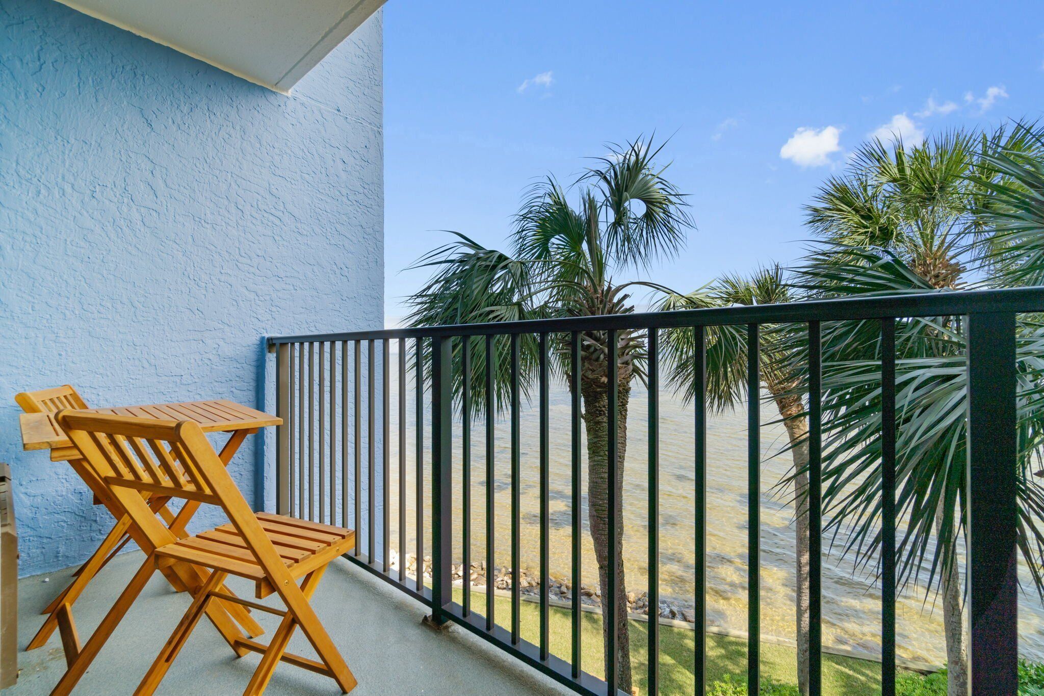 Un balcón con pared azul, sillas de madera y vistas a palmeras y al agua.