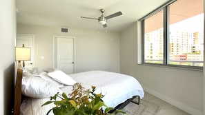 A bedroom with a bed, a ceiling fan, a window with a view of buildings, and a door.