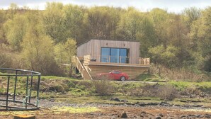 A small wooden cabin with a balcony, a red car parked in front, and a natural landscape with trees and shrubs.