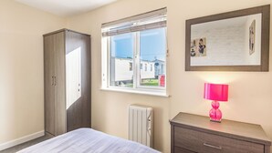 A bedroom with a bed, a wardrobe, a radiator, a window with a view of houses, and a bedside lamp. - Rainbow Bungalow 1 - UK39809 (Chapel St Leonards)