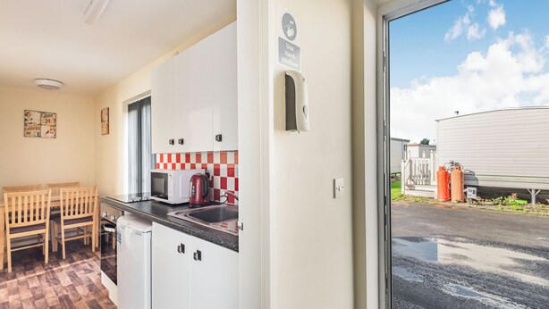 A kitchen with white cabinets, a microwave, a kettle, and a sink. There is a sign indicating the use of hand sanitizer. - Rainbow Bungalow 1 - UK39809 (Chapel St Leonards)