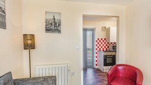A living room with a grey sofa, a red armchair, a radiator, and a kitchen visible through an open door. - Rainbow Bungalow 1 - UK39809 (Chapel St Leonards)