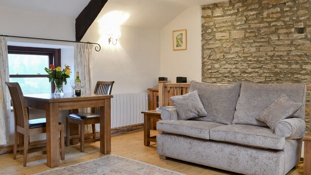 Una sala de estar acogedora con pared de piedra, un sofá gris, muebles de madera y un comedor con mesa y sillas.