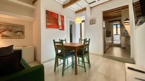 Un coin repas avec une table et des chaises en bois, un tableau accroché au mur et une cuisine visible à travers une porte ouverte.