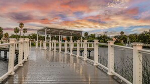 A wooden boardwalk with white railings, leading to a covered pavilion with a roof, surrounded by palm trees and a body of water. - East Riverside Downtown Bungalow (Port Richey)