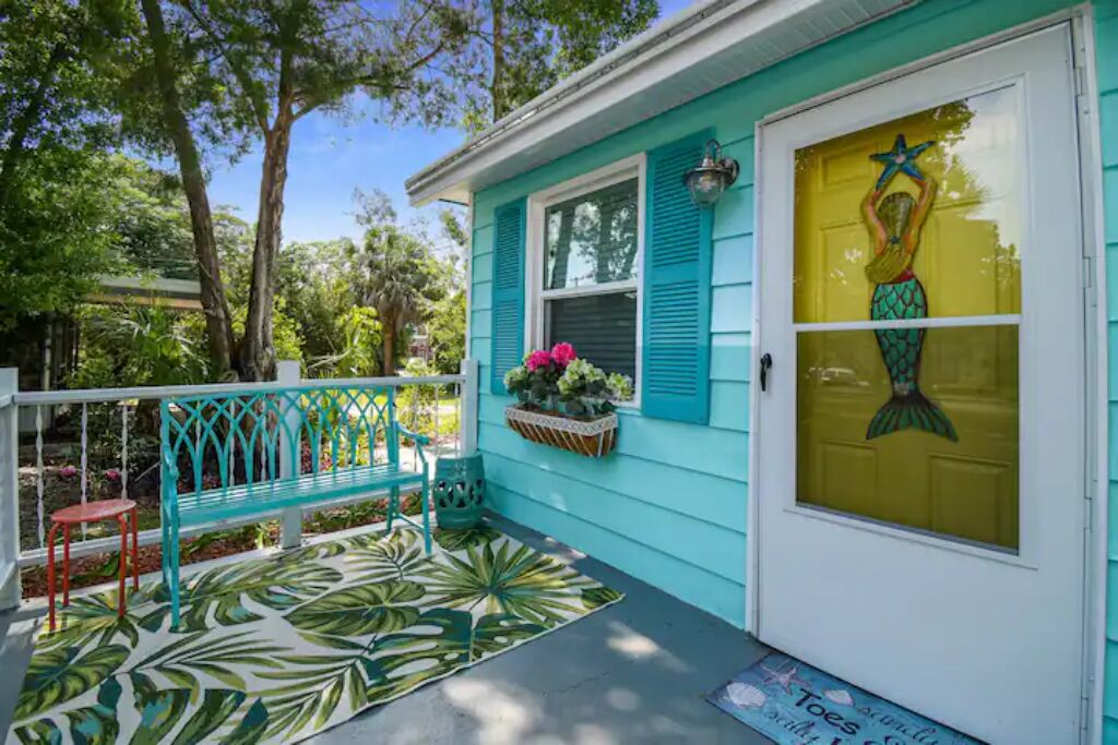 A turquoise house with a mermaid mural on the front door, a bench, and a tropical leaf-patterned rug.