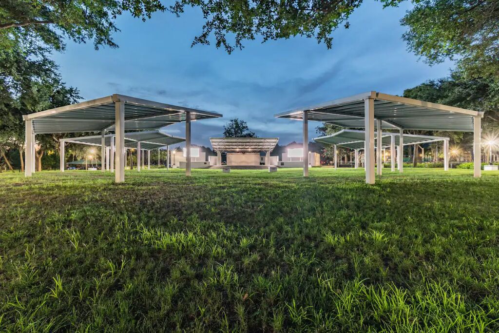 An open pavilion structure with a central seating area, surrounded by grass and trees.