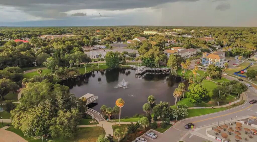 An aerial view of a park with a lake, a bridge, and surrounding buildings.