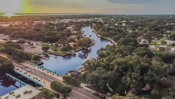 An aerial view of a riverside town with a bridge, roads, and greenery. - East Riverside Downtown Bungalow (Port Richey)