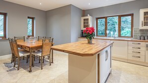 Een moderne keuken met een houten eettafel, rieten stoelen, een centraal kookeiland met een bloevas en grote ramen met uitzicht op bomen.