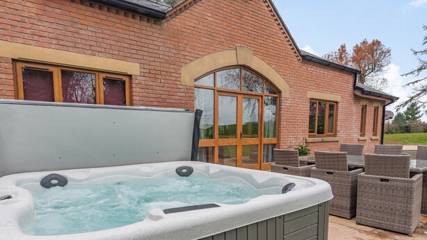 Een hottub met een houten terras en rieten meubilair voor een bakstenen huis.