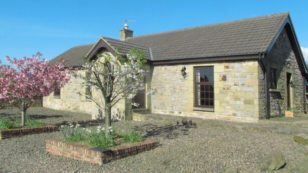 A stone building with a sloped roof, a chimney, and a small garden with flowering plants. - Robin Cottage Nr Rothbury (Weldon Bridge, near Rothbury)