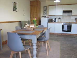 A dining area with a wooden table, grey chairs, a vase with flowers, and a kitchen in the background.