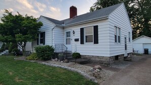 A two-story white house with black shutters, a front porch, and a small front yard with a tree and a rock garden. - Great Location - 3 miles to Cedar Point Park, 2.8 miles to CP Sports  Force Park (Sandusky)