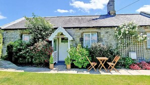 A stone cottage with a small porch, a chimney, and a garden with potted plants and a table with chairs.