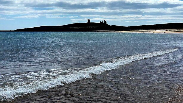 A beach with calm waters, a distant hill with structures, and a clear sky.