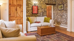 A cozy living room with a stone wall, a white sofa with green and red cushions, a wooden coffee table, and a chandelier.