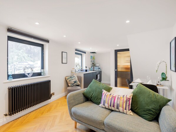 A modern bedroom with a grey sofa, a radiator, a window with a view of parked cars, and a kitchenette in the background.
