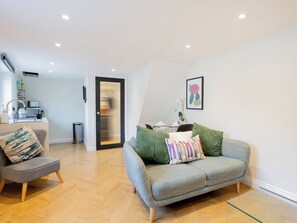 A modern living room with a grey sofa, a grey armchair, and a glass coffee table. There is a kitchen area with a sink and a refrigerator in the background.