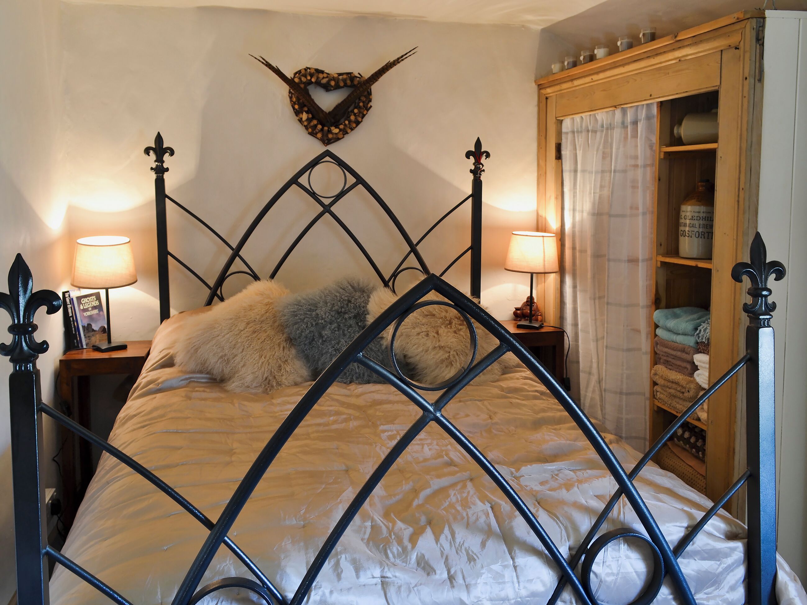 A bed with a diamond-patterned headboard, two bedside lamps, a heart-shaped wall decoration, and a wooden cabinet with towels.