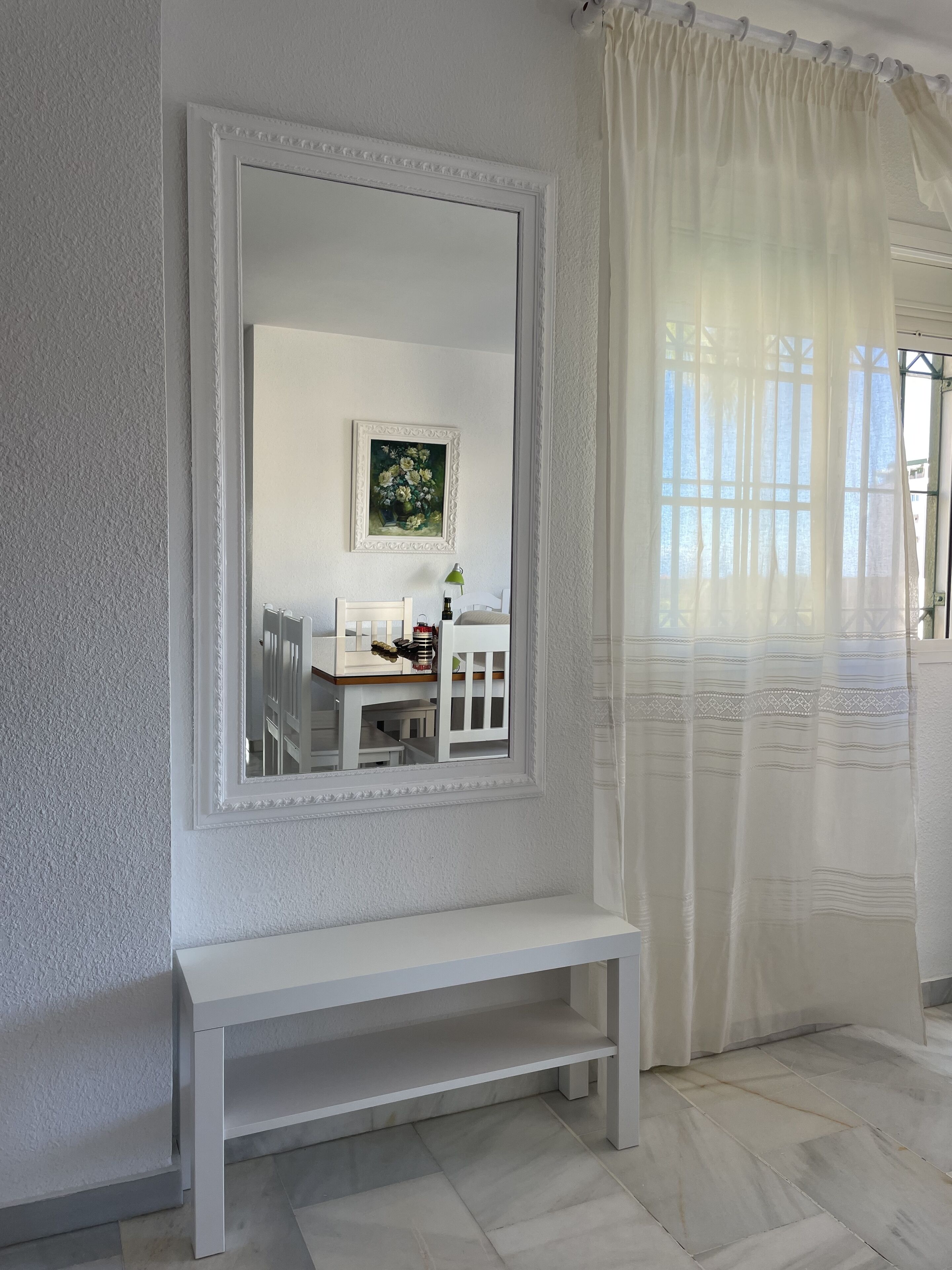 A room with a large mirror, a bench, a small table with a lamp, and a window with sheer curtains.