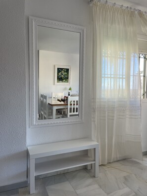Um quarto com um espelho grande, um banco, uma mesa pequena com uma luminária e uma janela com cortinas leves.
