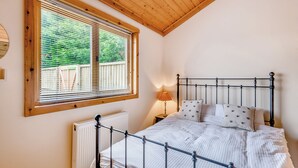Un dormitorio con techo de madera, una ventana con persianas, un radiador, estructura de cama metálica y una lámpara de noche.