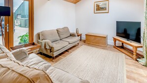 Una sala de estar acogedora, con techo de madera, un sofá beige, un televisor de pantalla plana sobre un mueble de madera y un ventanal con vistas a un estacionamiento.