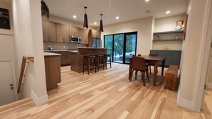 Een moderne keuken met houten kastjes, een centraal eiland met krukken, een eettafel met stoelen en een groot raam met uitzicht op bomen.