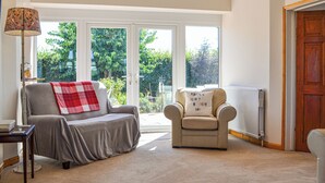 A living room with a sofa, armchair, and a lamp. There is a window with a view of greenery outside. - 4 bedroom accommodation in Hunstanton (Hunstanton)